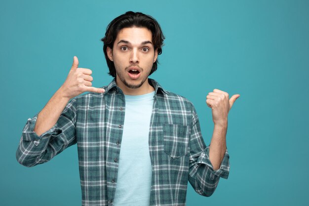 surprised young handsome man looking at camera showing call gesture pointing to side isolated on blue background