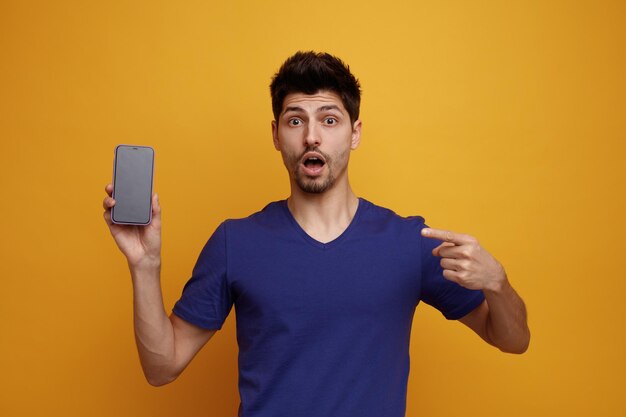 Surprised young handsome man looking at camera showing mobile phone to camera pointing to mobile phone on yellow background