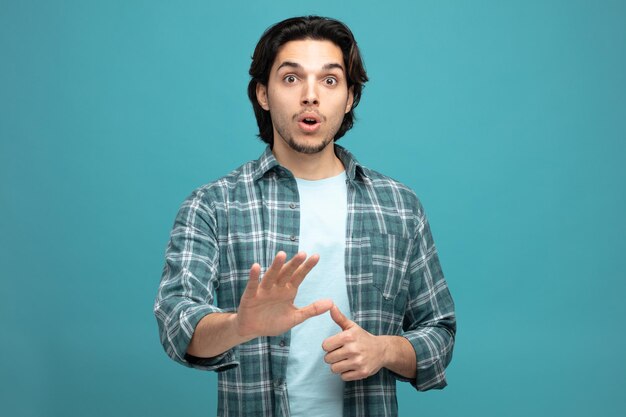 surprised young handsome man looking at camera showing thumb up and no gesture isolated on blue background