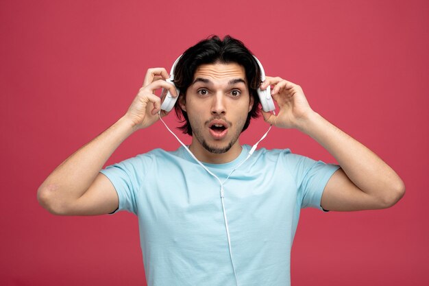 surprised young handsome man looking at camera taking headphones off isolated on red background