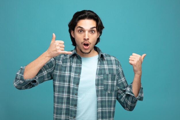 surprised young handsome man showing call me gesture looking at camera pointing to side isolated on blue background