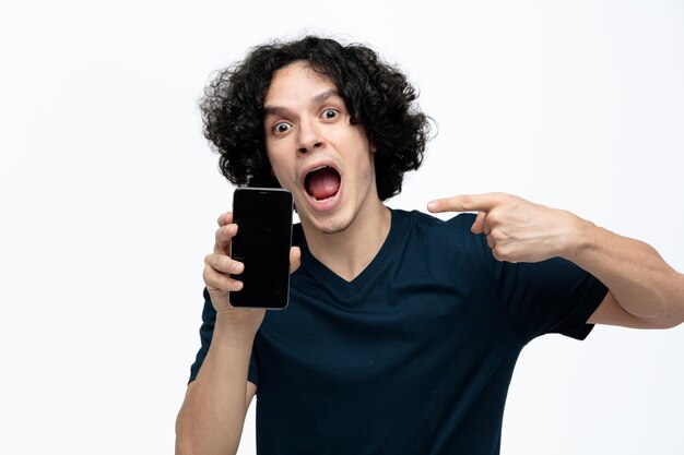 Удивленный молодой красивый мужчина показывает мобильный телефон камере, указывая на него, глядя на камеру, изолированную на белом фоне