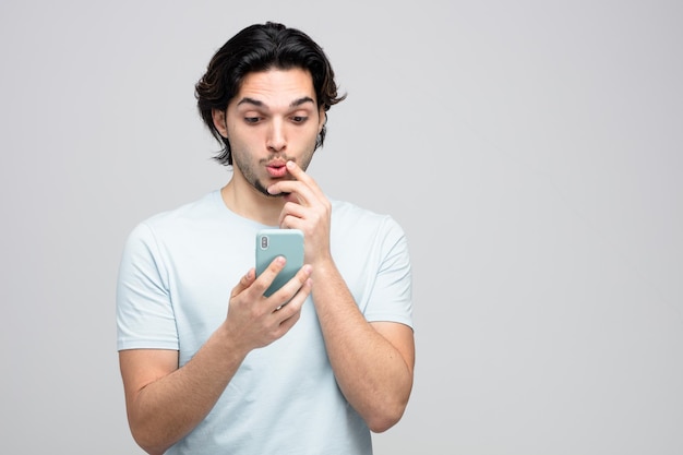удивленный молодой красивый мужчина трогает подбородок, держа и глядя на мобильный телефон, изолированный на белом фоне с копировальным пространством