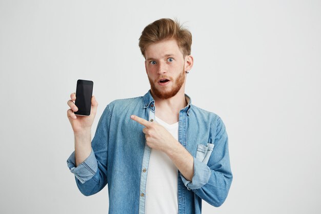 Surprised young handsome man with opened mouth pointing finger at phone in his hand.