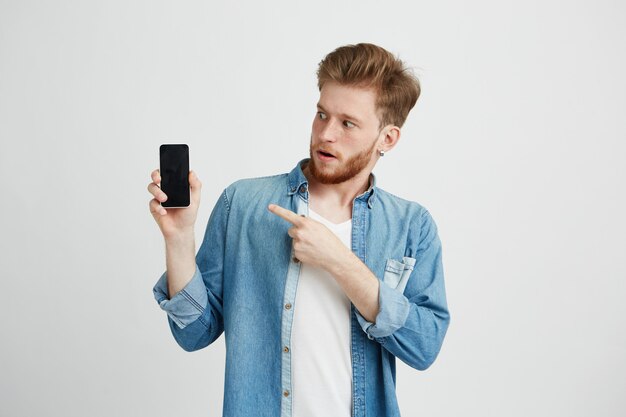 Surprised young handsome man with opened mouth pointing finger at phone in his hand.