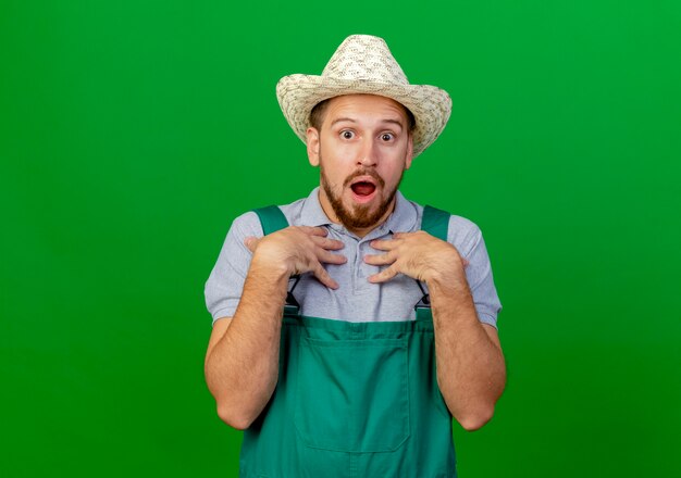 Surprised young handsome slavic gardener in uniform and hat looking keeping hands on chest 