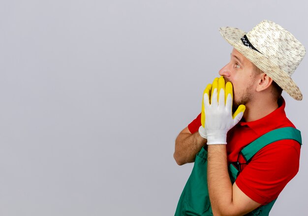 Удивленный молодой красивый славянский садовник в униформе и шляпе в перчатках садовника, стоящий в профиль, держа руки во рту, глядя прямо изолированно