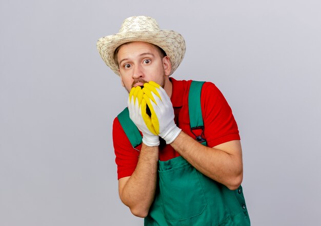 Surprised young handsome slavic gardener in uniform wearing gardening gloves and hat looking keeping hands on mouth isolated