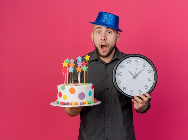 Surprised young handsome slavic party guy wearing party hat holding birthday cake with stars and clock looking at camera isolated on crimson background with copy space