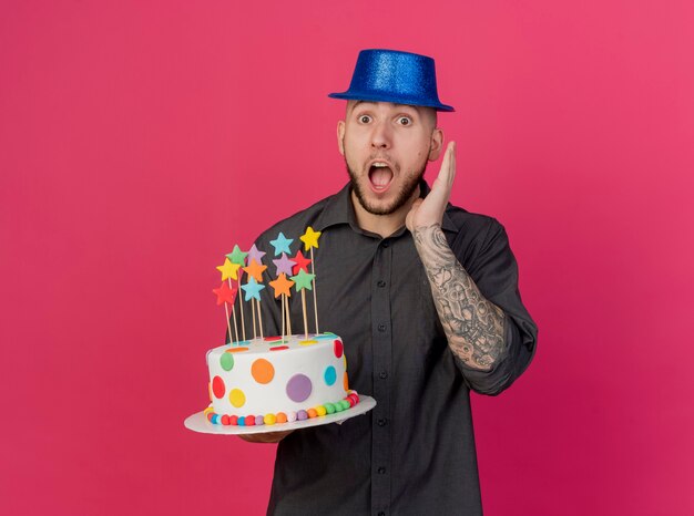 Surprised young handsome slavic party guy wearing party hat holding birthday cake with stars keeping hand in air looking at camera isolated on crimson background with copy space