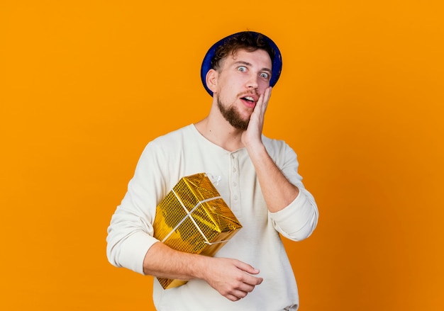 Free photo surprised young handsome slavic party guy wearing party hat holding gift box looking at camera touching face isolated on orange background with copy space