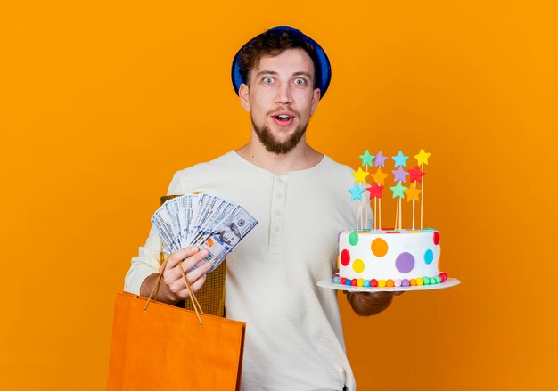 Surprised young handsome slavic party guy wearing party hat holding gift box money paper bag and birthday cake with stars looking at camera isolated on orange background