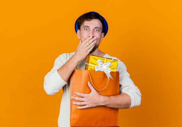 Surprised young handsome slavic party guy wearing party hat holding gift boxes in paper bag looking at camera keeping hand on mouth isolated on orange background with copy space