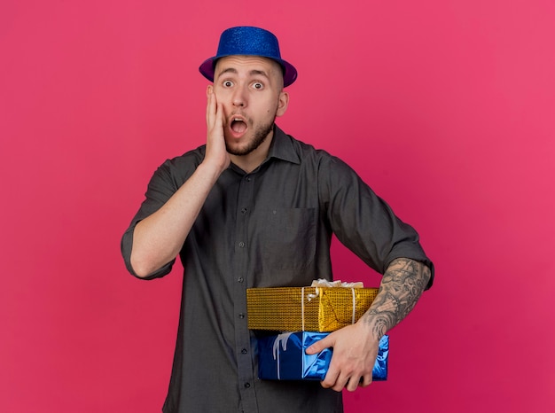 Surprised young handsome slavic party guy wearing party hat holding gift packs keeping hand on face looking at camera isolated on crimson background with copy space