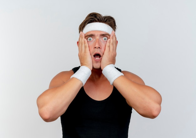 Surprised young handsome sporty man wearing headband and wristbands putting hands on face isolated on white wall