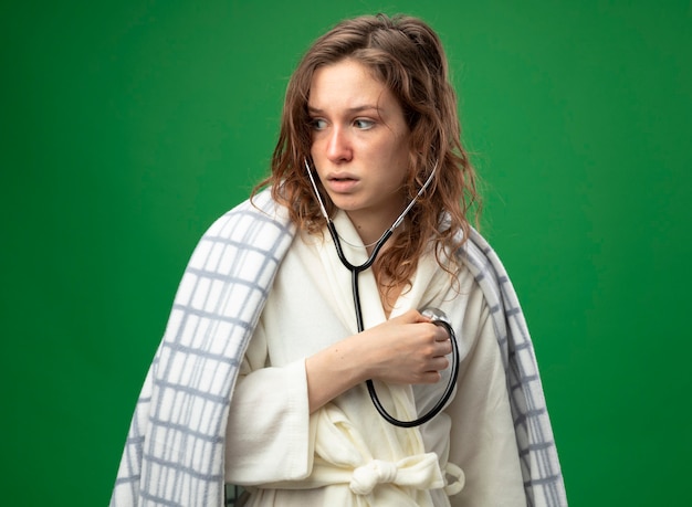 Surprised young ill girl looking at side wearing white robe wrapped in plaid listening to her own heartbeat isolated on green