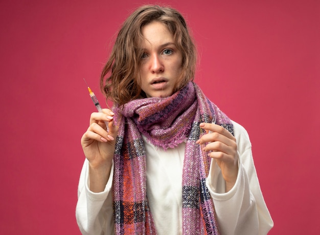 Foto gratuita giovane ragazza malata sorpresa che osserva la siringa della tenuta della sciarpa e della veste bianca da portare con la fiala isolata sulla parete rosa