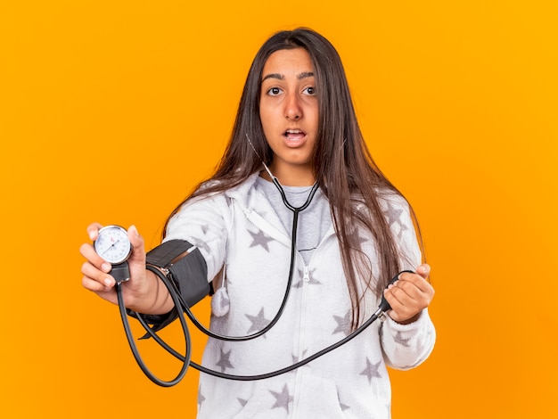 Free photo surprised young ill girl measuring her own pressure with sphygmomanometer isolated on yellow background
