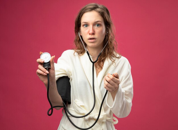 Surprised young ill girl wearing white robe measuring her own pressure with sphygmomanometer isolated on pink