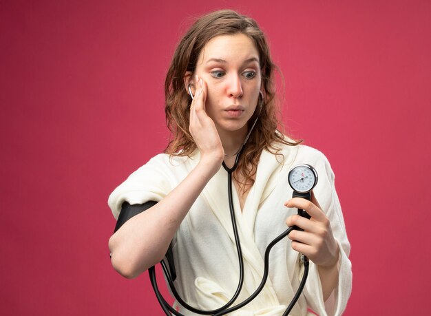 Удивленная молодая больная девушка в белом халате измеряет собственное давление с помощью сфигмоманометра, положив руку на щеку, изолированную на розовом