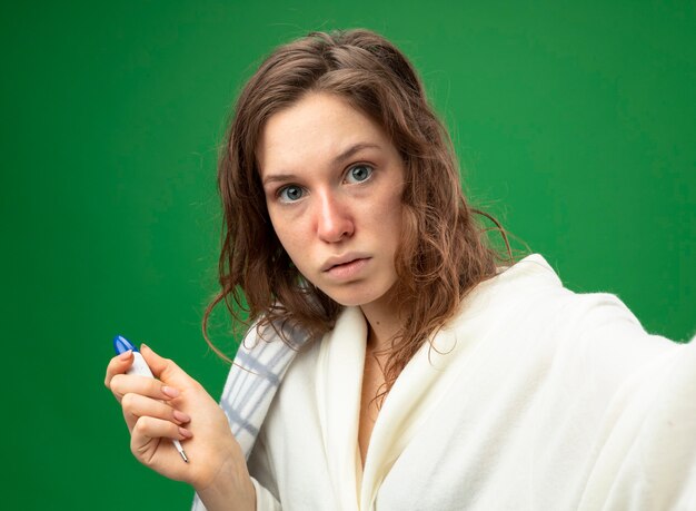 Surprised young ill girl wearing white robe wrapped in plaid holding thermometer and camera isolated on green
