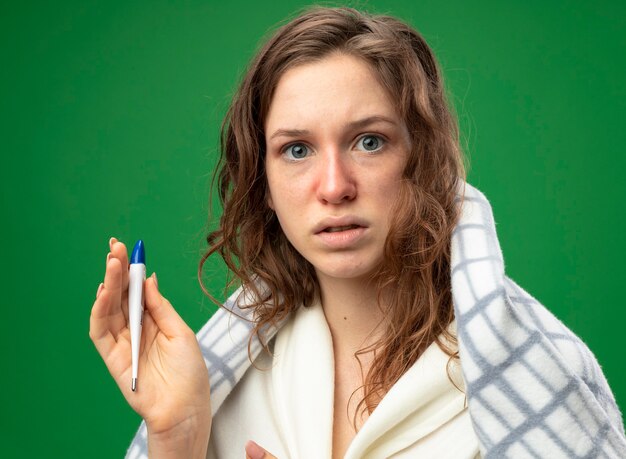 Удивленная молодая больная девушка в белом халате, завернутая в плед, с термометром, изолированным на зеленом