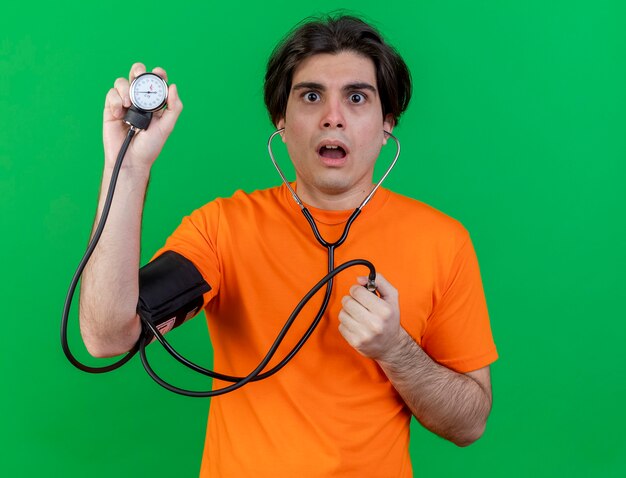 Surprised young ill man measuring his own pressure with sphygmomanometer isolated on green