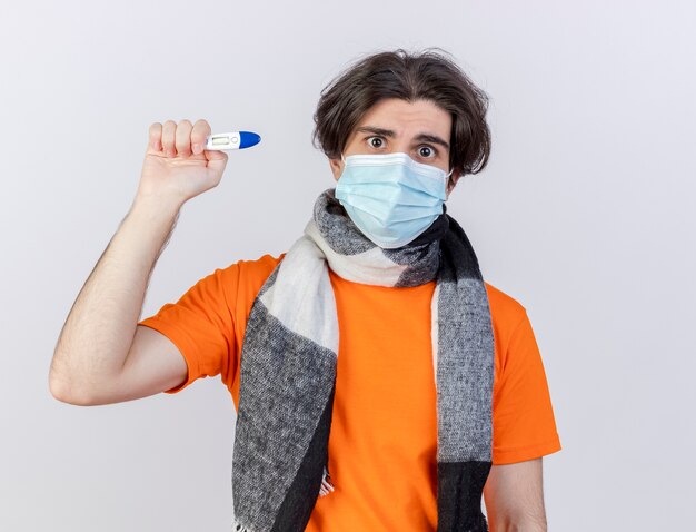 Удивленный молодой больной в шарфе и медицинской маске, держащий термометр на белом фоне