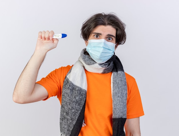 Surprised young ill man wearing scarf and medical mask holding thermometer isolated on white background