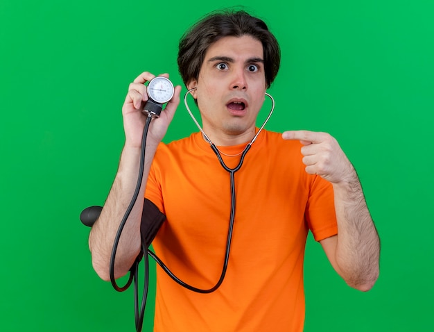 Surprised young ill man wearing stethoscope holding and points at sphygmomanometer isolated on green