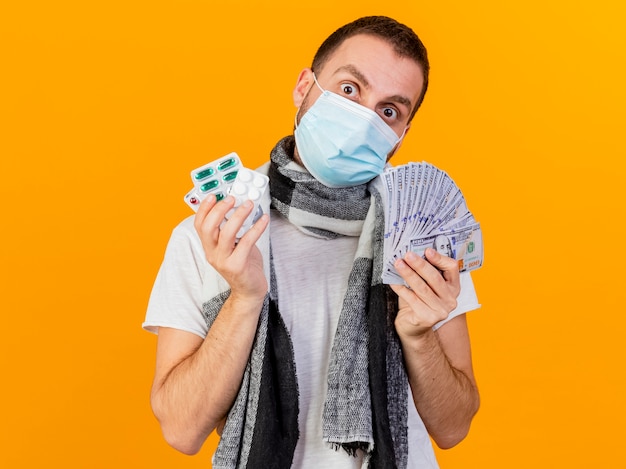 Удивленный молодой больной в зимней шапке и медицинской маске держит деньги с таблетками, изолированными на желтом фоне