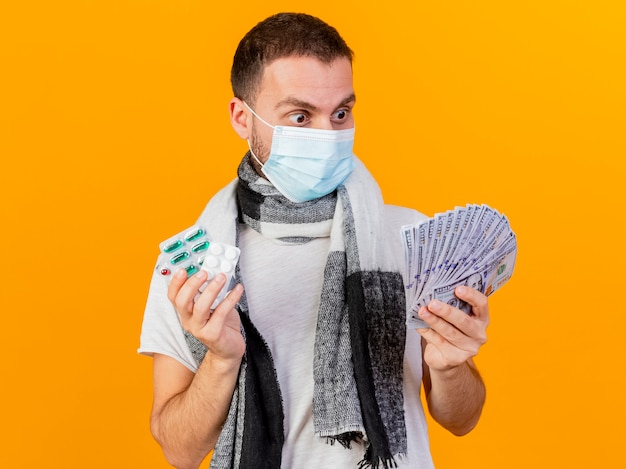 Удивленный молодой больной в зимней шапке и медицинской маске держит таблетки и смотрит на деньги в руке, изолированной на желтом фоне