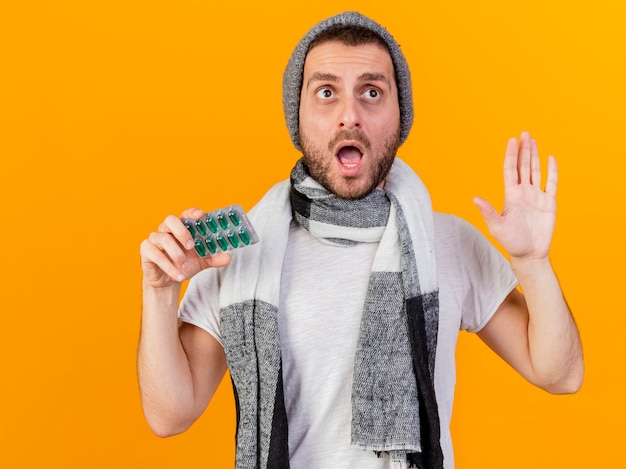 Free photo surprised young ill man wearing winter hat and scarf holding pills and showing stop gesture isolated on yellow background