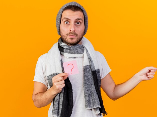 Surprised young ill man wearing winter hat and scarf holding question mark note and points at side isolated on yellow background