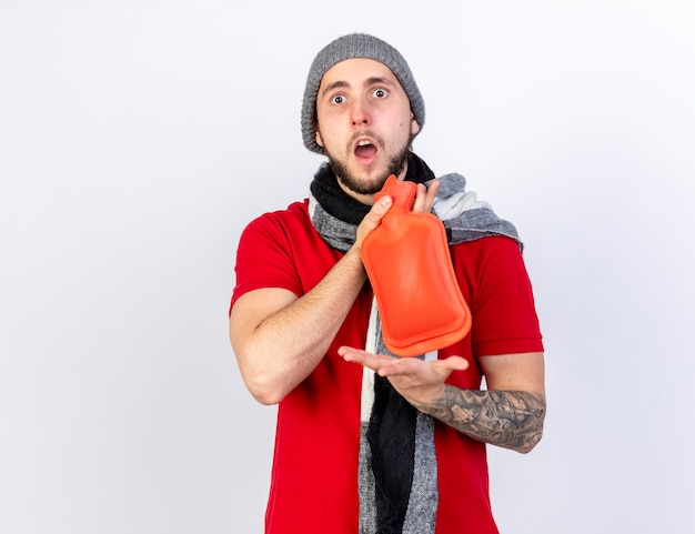 Surprised young ill man wearing winter hat and scarf holds hot water bottle isolated on white wall