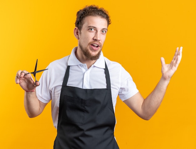 Surprised young male barber wearing uniform holding scissors isolated on yellow background