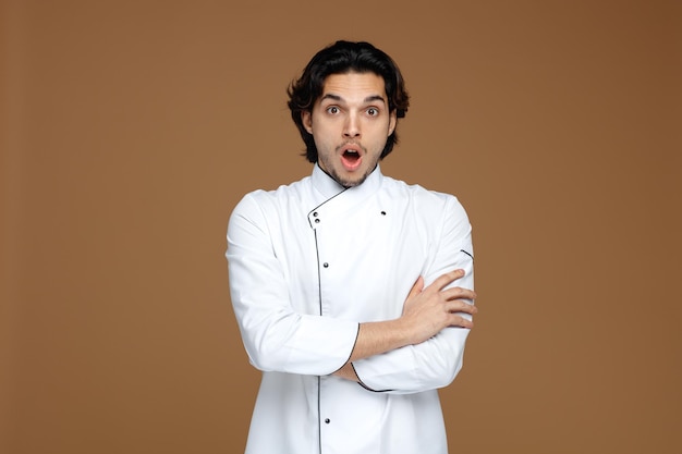 surprised young male chef wearing uniform keeping arms crossed looking at camera isolated on brown background