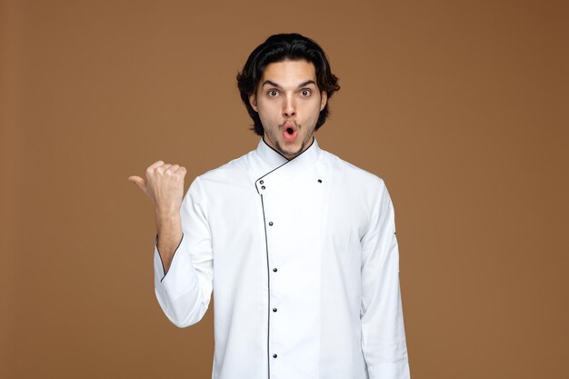 surprised young male chef wearing uniform looking at camera pointing to side isolated on brown background