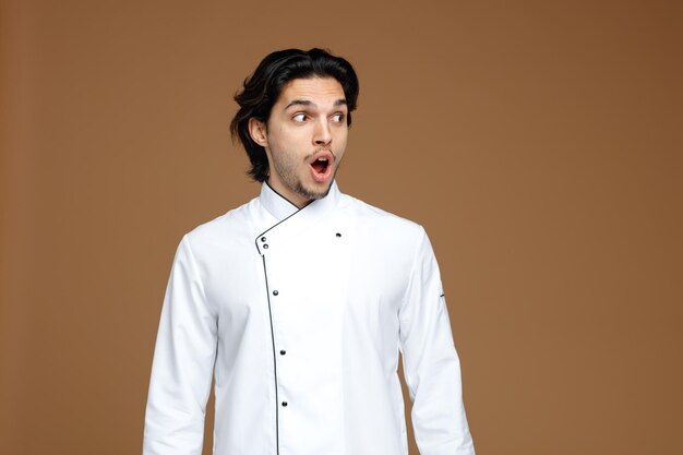 surprised young male chef wearing uniform looking at side isolated on brown background