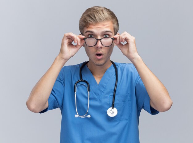 Surprised young male doctor wearing doctor uniform with stethoscope and glasses isolated on white wall