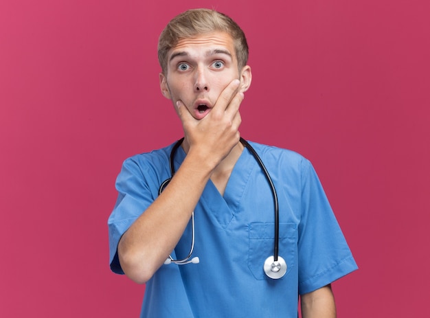 청진기와 의사 유니폼을 입고 놀란 젊은 남성 의사 핑크 벽에 고립 된 턱을 잡고