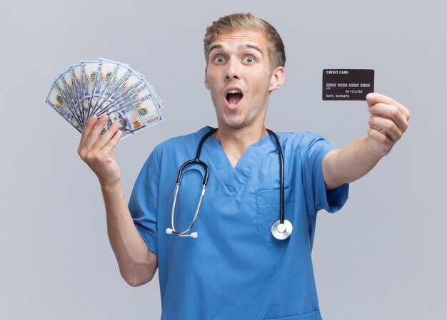 Surprised young male doctor wearing doctor uniform with stethoscope holding out cash and credit card  isolated on white wall