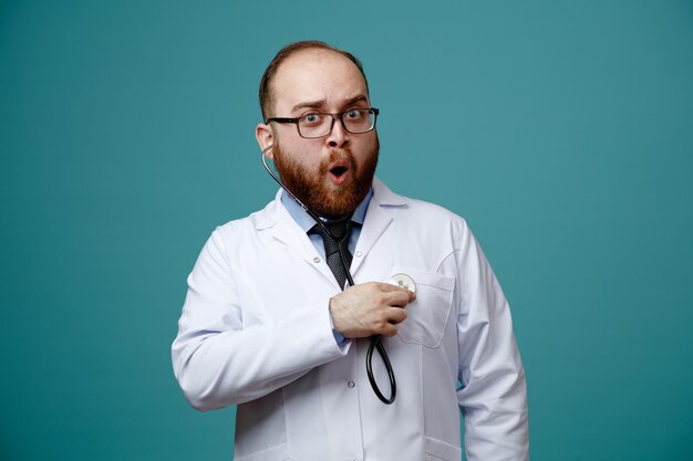 surprised young male doctor wearing glasses medical coat and stethoscope looking at camera listening to his heartbeats with stethoscope isolated on blue background