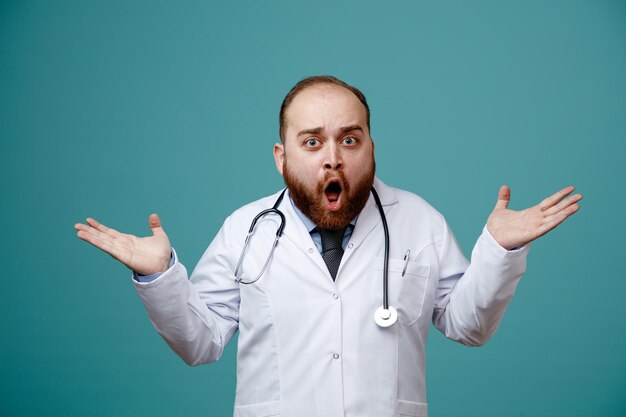 Surprised young male doctor wearing medical coat and stethoscope around his neck looking at camera showing empty hands isolated on blue background