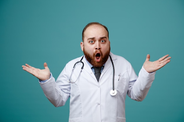 Удивленный молодой врач-мужчина в медицинском халате и стетоскопе на шее смотрит в камеру, показывающую пустые руки, изолированные на синем фоне