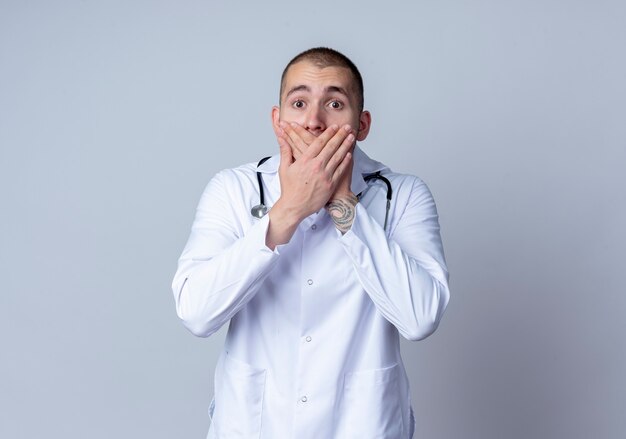 Surprised young male doctor wearing medical robe and stethoscope around his neck covering mouth with hands isolated on white wall