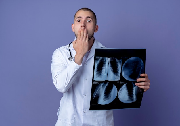 Surprised young male doctor wearing medical robe and stethoscope around his neck holding x-ray shot and putting hand on mouth isolated on purple wall
