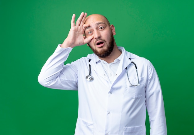 Surprised young male doctor wearing medical robe and stethoscope opening eye with hand isolated on green