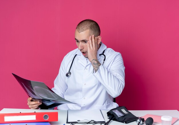 Surprised young male doctor wearing medical robe and stethoscope sitting at desk with work tools holding and looking at x-ray shot with hand on head isolated on pink wall