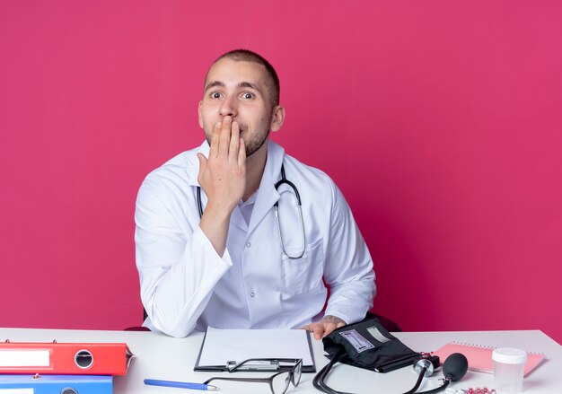 Surprised young male doctor wearing medical robe and stethoscope sitting at desk with work tools putting hand on mouth isolated on pink wall